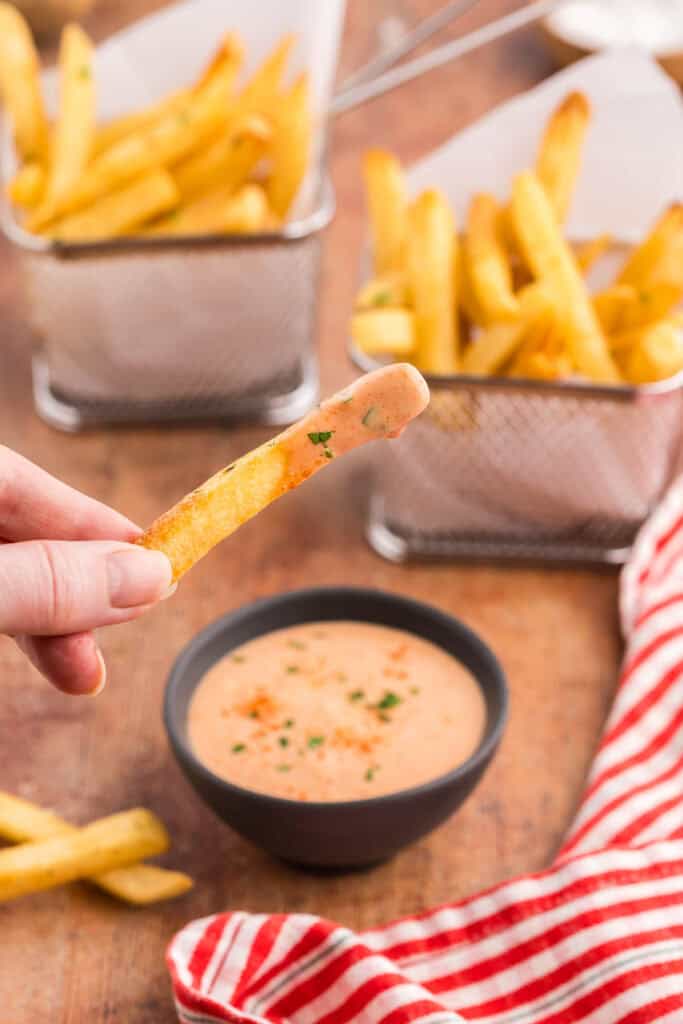 Close up shot of a golden brown french fry just having been dipped into a creamy orangey/pink Fry Sauce.  In the background are two small baskets of Air Fryer frozen french fries.