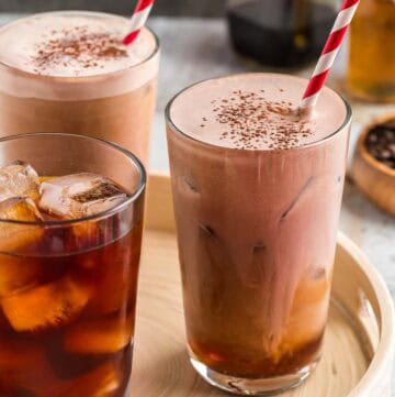 Three large glasses on a serving tray. Two of them are filled with chocolate cream cold brew with red and white striped straws and cocoa sprinkled on top. One glass only has ice and cold brew coffee in it, waiting to have the chocolate cream added.