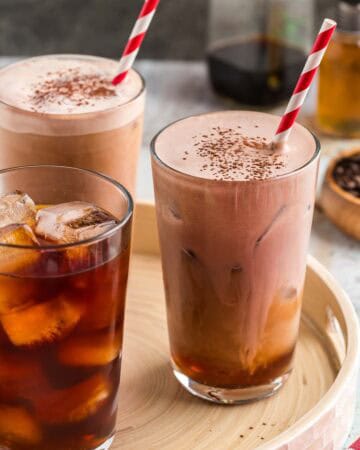 Three large glasses on a serving tray. Two of them are filled with chocolate cream cold brew with red and white striped straws and cocoa sprinkled on top. One glass only has ice and cold brew coffee in it, waiting to have the chocolate cream added.