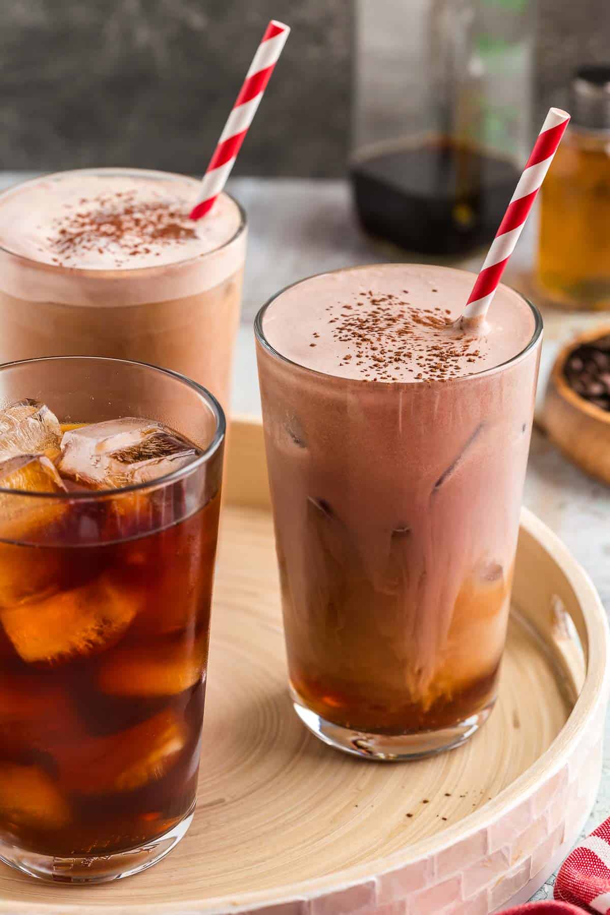 Three large glasses on a serving tray. Two of them are filled with chocolate cream cold brew with red and white striped straws and cocoa sprinkled on top. One glass only has ice and cold brew coffee in it, waiting to have the chocolate cream added.