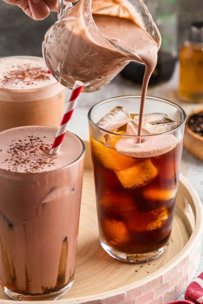 Three drinks sitting on a serving tray. Two of them are filled with creamy brown chocolate cream cold brew with cocoa sprinkled on top and have a red and white striped straw. The third tall glass is filled with ice and dark cold brew coffee. A small glass jug is pouring in chocolate cream on top.