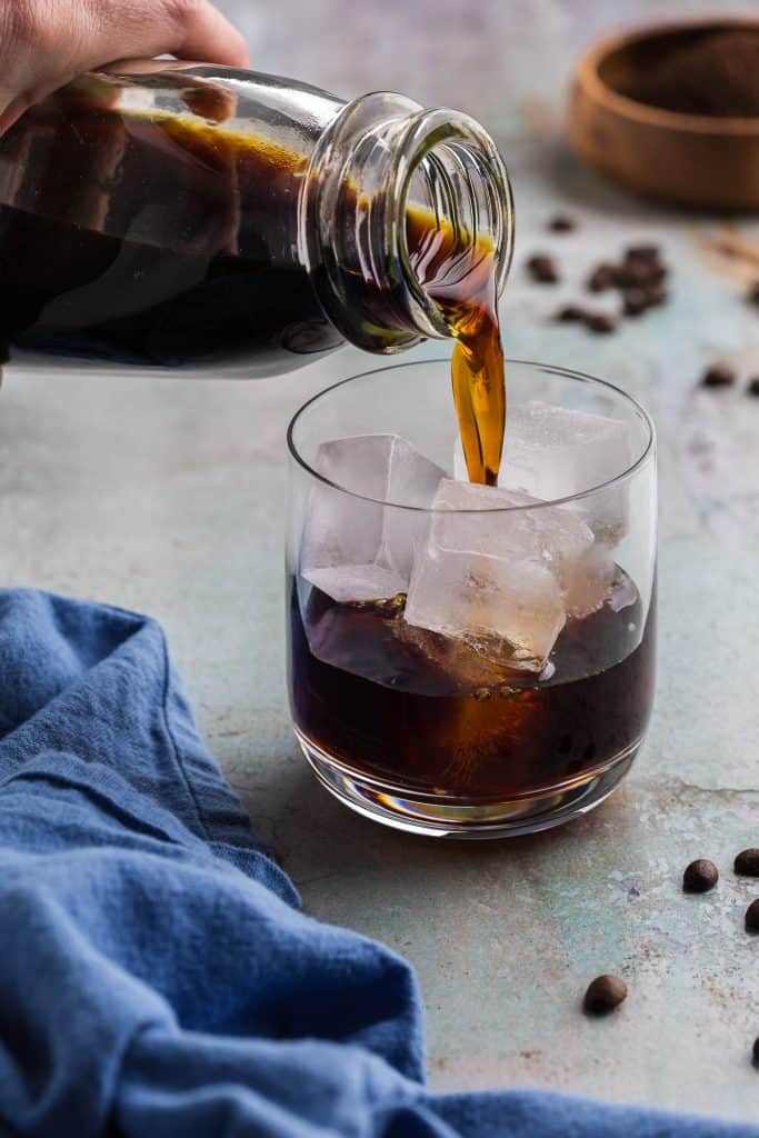 side shot of a low ball glass filled with ice and cold brew concentrate being poured in. The concentrate is a dark liquid with amber highlights where the light catches it on the ice cubes.