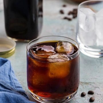 A low ball glass of cold brew and ice is in the centre of the shot. The cold brew is a dark liquid with amber highlights where the lighting is coming through. In the background is a container of cold brew concentrate, as well as a small wooden bowl filled with coffee beans. Coffee beans are also scattered around the scene.