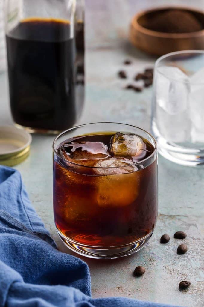 A low ball glass of cold brew and ice is in the centre of the shot. The cold brew is a dark liquid with amber highlights where the lighting is coming through. In the background is a container of cold brew concentrate, as well as a small wooden bowl filled with coffee beans. Coffee beans are also scattered around the scene.