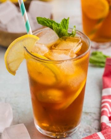 Up close side shot of a pint glass filled with ice and cold brew tea. It is a golden brown color and there are two lemon wheels floating between the ice and the side of the glass. The drink is garnished with half a lemon wheel hanging off the lip of the glass and a sprig of fresh mint. There is a light blue and white straw.