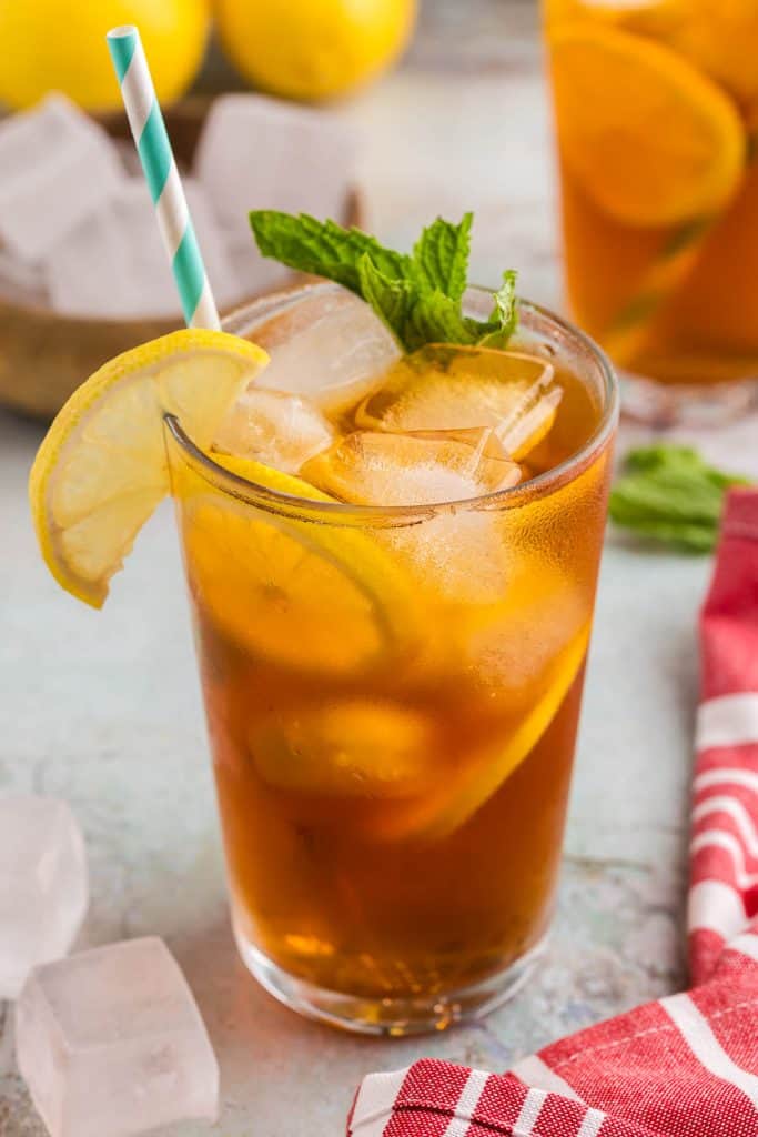 Up close side shot of a pint glass filled with ice and cold brew tea. It is a golden brown color and there are two lemon wheels floating between the ice and the side of the glass. The drink is garnished with half a lemon wheel hanging off the lip of the glass and a sprig of fresh mint. There is a light blue and white straw.