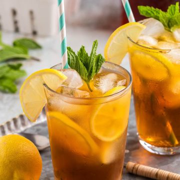 Side shot of two pint glasses on a silver serving tray. Each glass is filled with ice and cold brew tea. Each glass has lemon wheels floating between the ice and the side of the glass, and each are garnished with fresh mint and half a lemon wheel on the edge of the rim. Each also have a light blue and white straw. A basket of lemons are in the background.
