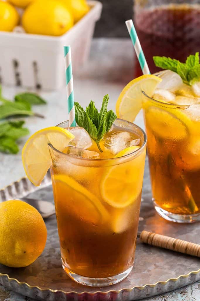 Side shot of two pint glasses on a silver serving tray. Each glass is filled with ice and cold brew tea. Each glass has lemon wheels floating between the ice and the side of the glass, and each are garnished with fresh mint and half a lemon wheel on the edge of the rim. Each also have a light blue and white straw. A basket of lemons are in the background.