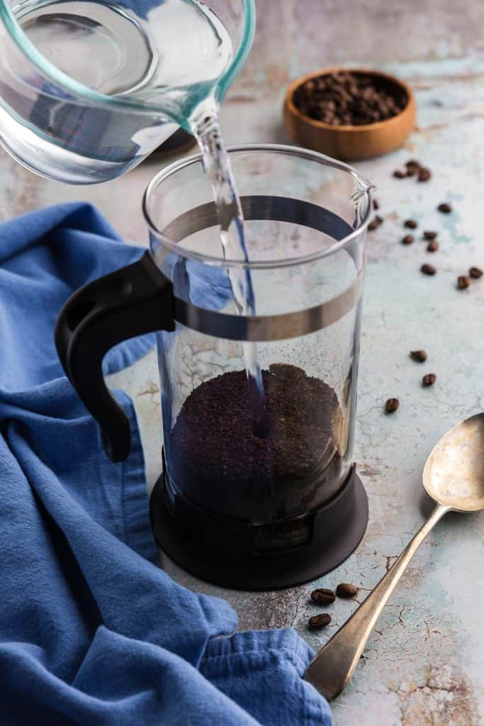 45 degree angle shot of a French press 1/4 filled with coarsely ground coffee. Water is being poured in from above. There is a spoon lying next to the French press and coffee beans are scattered around the scene. This will become the cold brew concentrate.