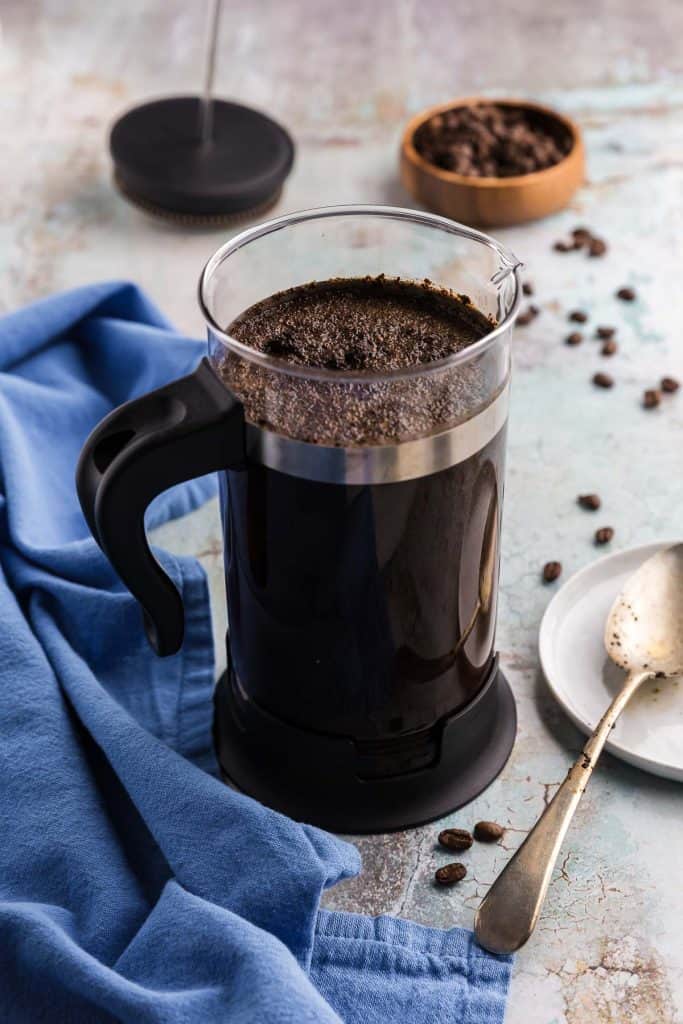 45 degree angle shot of a French press filled with water and coarsely ground coffee. A lot of the coarsely ground coffee is floating at the surface. A spoon which was used to stir the coffee mixture is resting next to the French press.