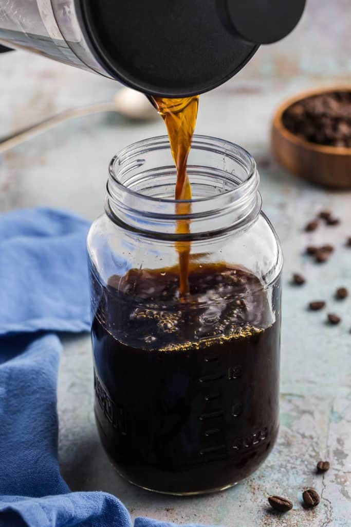 Pouring our the cold brew concentrate that was brewing in a French press into a mason jar. The mason jar is 2/3rd full of cold brew concentrate and a stream of concentrate is still flowing out of the French press. Coffee beans are scattered around the scene.