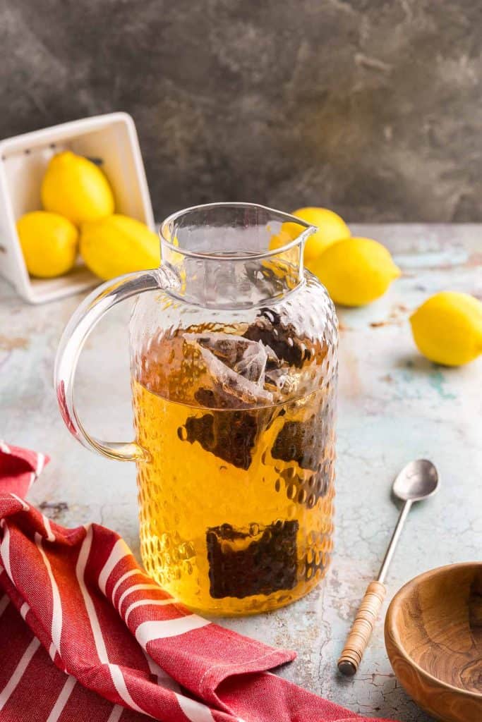Beginning the brewing process for cold brew tea. A large pitcher of water with 8 tea bags recently added in. The color of the water has turned slightly brown as the tea begins to brew. In the background a container of lemons is on its side and lemons have spilled out. A long spoon is laying beside the pitcher, and a red linen lies next to the pitcher.