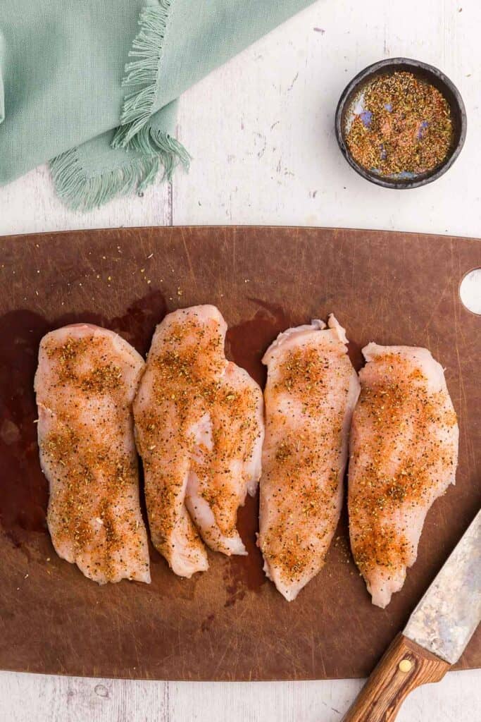 Overhead shot of chicken breasts for lemon chicken orzo pasta cut in half horizontally and seasoned. The chicken breasts are on a wooden cutting board and have plenty of seasoning which is red from the paprika and flakes of oregano are apparent.