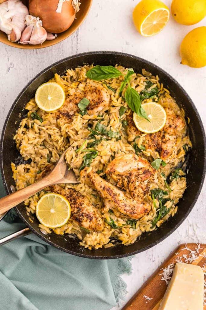 Overhead shot with a frypan in the middle filled with lemon chicken orzo pasta. The chicken is a golden brown and surrounded by a yellowy/brown orzo in sauce. Vibrant green spinach accents the dish. Three bright yellow lemon wheel slices are in the pan. In the upper left corner of the scene is a wooden bowl filled with fresh onion and garlic. Fresh lemons are in the upper right corner and the bottom right corner is a wedge of parmesan cheese on a wooden cutting board.