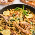 45 degree angle shot of a black fry pan filled with fully cooked Lemon Chicken Orzo Pasta. 4 Seasoned golden brown chicken breasts sit in a fry pan filled with creamy lemon orzo garnished with torn basil leaves and lemon slices. In the background is a wooden bowl of fresh onion and garlic and a wedge of parmesan cheese.