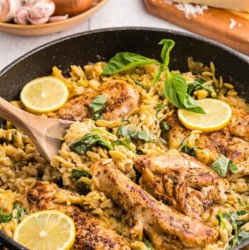 45 degree angle shot of a black fry pan filled with fully cooked Lemon Chicken Orzo Pasta. 4 Seasoned golden brown chicken breasts sit in a fry pan filled with creamy lemon orzo garnished with torn basil leaves and lemon slices. In the background is a wooden bowl of fresh onion and garlic and a wedge of parmesan cheese.