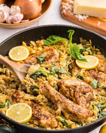 45 degree angle shot of a black fry pan filled with fully cooked Lemon Chicken Orzo Pasta. 4 Seasoned golden brown chicken breasts sit in a fry pan filled with creamy lemon orzo garnished with torn basil leaves and lemon slices. In the background is a wooden bowl of fresh onion and garlic and a wedge of parmesan cheese.