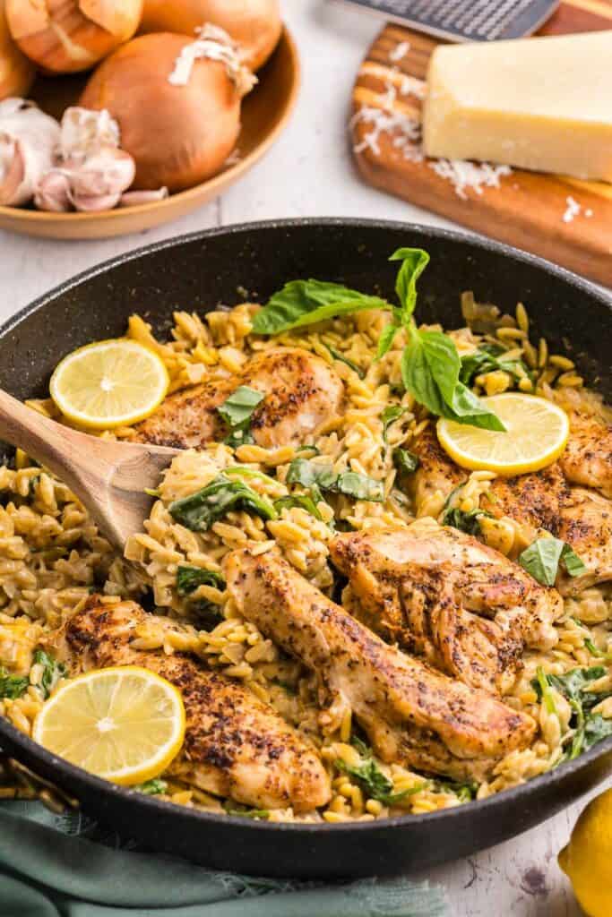 45 degree angle shot of a black fry pan filled with fully cooked Lemon Chicken Orzo Pasta. 4 Seasoned golden brown chicken breasts sit in a fry pan filled with creamy lemon orzo garnished with torn basil leaves and lemon slices. In the background is a wooden bowl of fresh onion and garlic and a wedge of parmesan cheese.