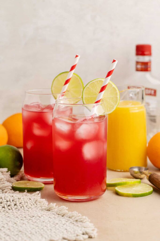 Two tall glasses are filled with a red drink called madras cocktail. The glasses are each garnished with a lime wheel perched on the lip of the glass, and each glass has a red and white striped straw. Both drinks are filled with with. In the background is a jug of orange juice, a bottle of vodka, and fresh oranges and limes.