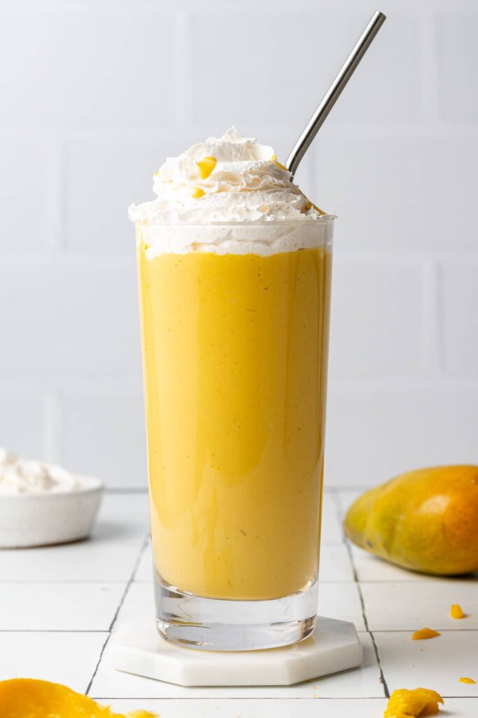 A close up side shot of a mango milkshake served in a highball glass. The bright orange milkshake is topped with thick whip cream and has a stainless steel straw.
