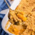 45 degree angle of a peach cobbler with cake mix after coming out of the oven. A serving spoon is lifting up a scoop filled with peaches and a crunchy topping.