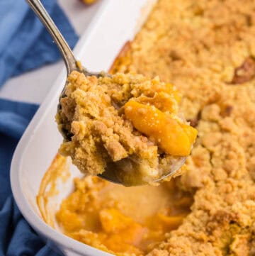45 degree angle of a peach cobbler with cake mix after coming out of the oven. A serving spoon is lifting up a scoop filled with peaches and a crunchy topping.