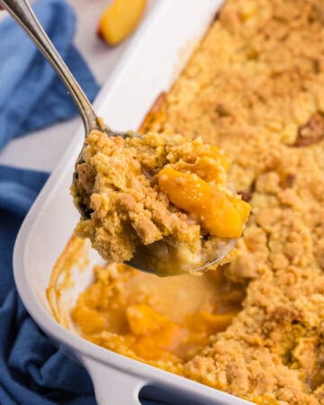 45 degree angle of a peach cobbler with cake mix after coming out of the oven. A serving spoon is lifting up a scoop filled with peaches and a crunchy topping.