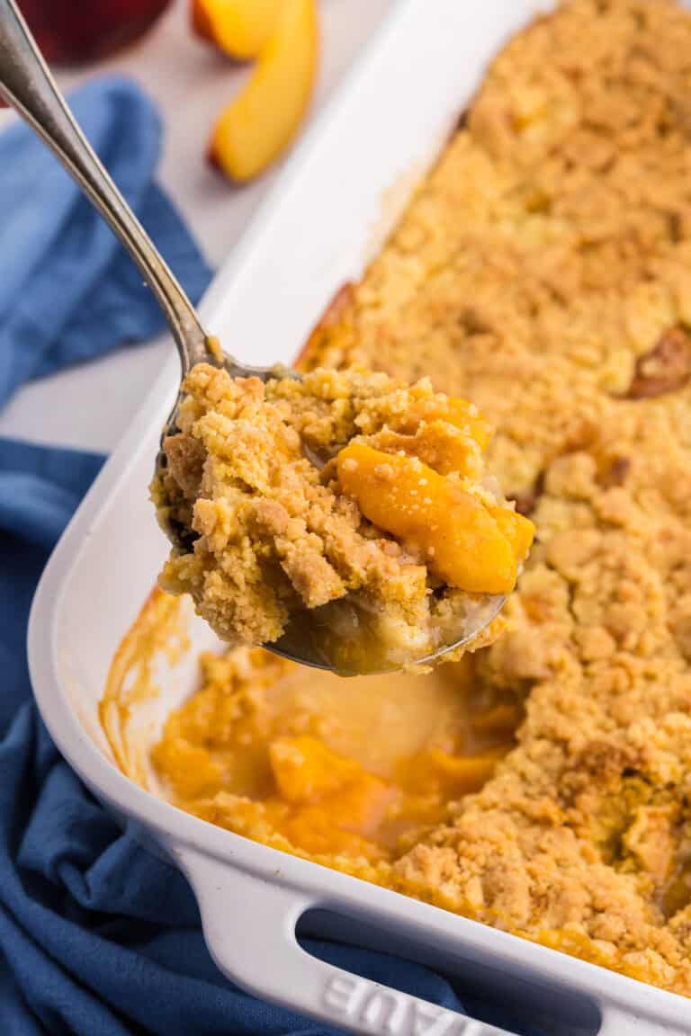 45 degree angle of a peach cobbler with cake mix after coming out of the oven. A serving spoon is lifting up a scoop filled with peaches and a crunchy topping.