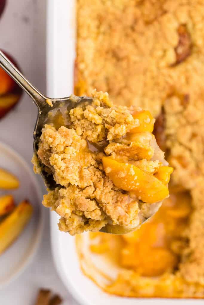 Overhead close up shot of a serving spoon filled with peach cobbler made with cake mix. Peach slices are evident and covered in a crunch topping. A gooey sauce is also evident on the spoon. Below the spoon and out of focus is a 9x13 white casserole dish filled with peach cobbler but missing a spoonful. Fresh peaches and peach slices are peeking in to the scene along the left hand side.