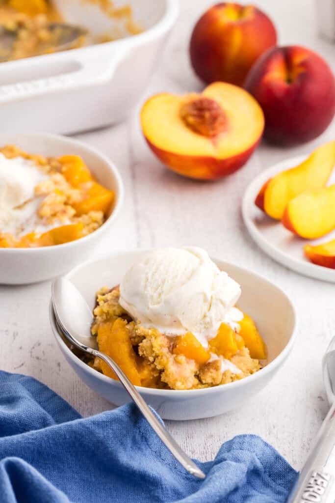 Peach cobbler with cake mix served in small white bowls with a scoop of ice cream. In the background are fresh pieces, and slices of pieces.