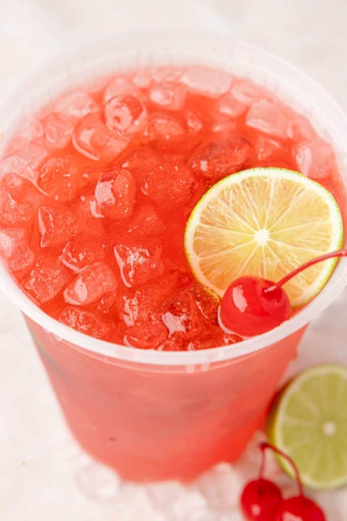 Close up shot looking down on a plastic glass filled with ice and bright red/pink cherry limeade. The drink is garnished with a lime slice as well as a maraschino cherry.