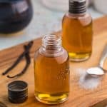 Side shot of two small glass bottles filled with a golden liquid of vanilla simple syrup. The bottles are on a wooden cutting board. The bottle closer to the camera has it's lid off and set beside the bottle on the left. There is also two vanilla beans on the left of the bottle and on the right is a teaspoon filled with white sugar.