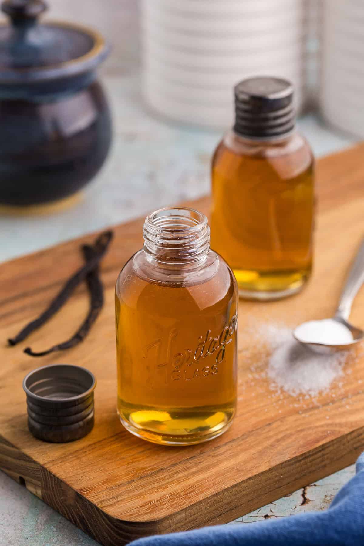 Side shot of two small glass bottles filled with a golden liquid of vanilla simple syrup. The bottles are on a wooden cutting board. The bottle closer to the camera has it's lid off and set beside the bottle on the left. There is also two vanilla beans on the left of the bottle and on the right is a teaspoon filled with white sugar.