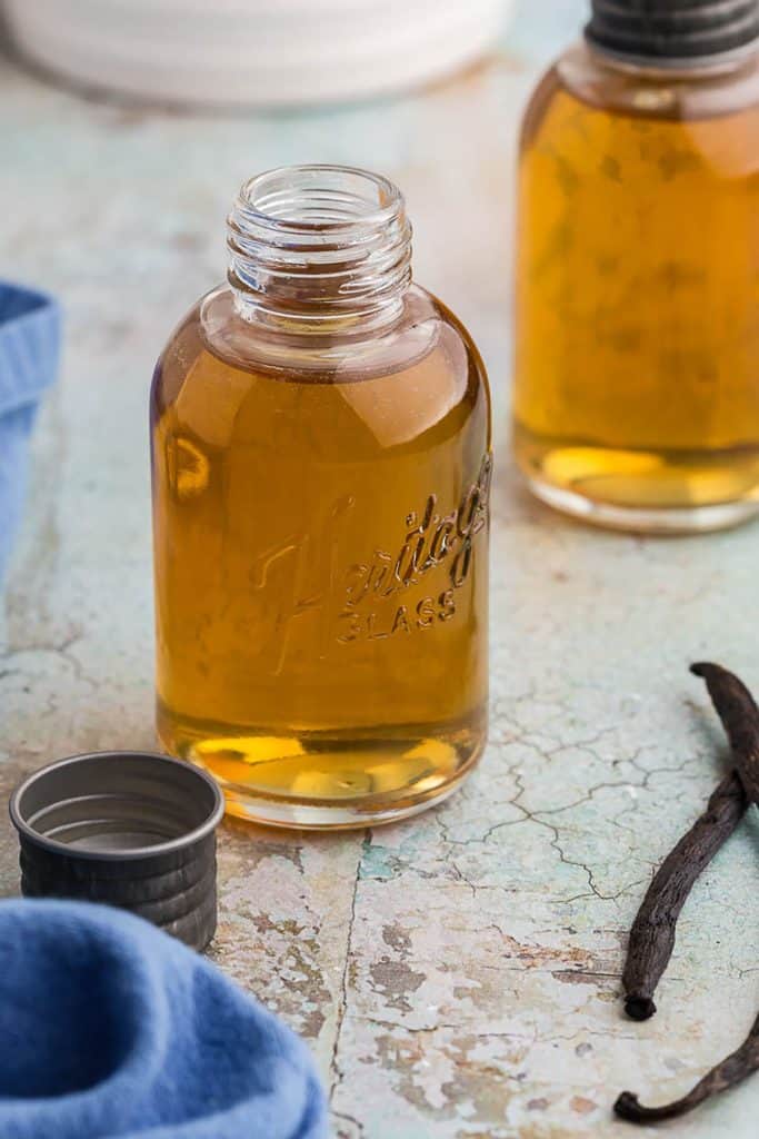 Side shot of a bottle of containing a golden hue of liquid known as vanilla simple syrup. The lid is off the bottle and sitting to the left of it. On the right and up a bit is another bottle of vanilla simple syrup, and between them are two vanilla beans which help to indicate the flavor of the syrup.