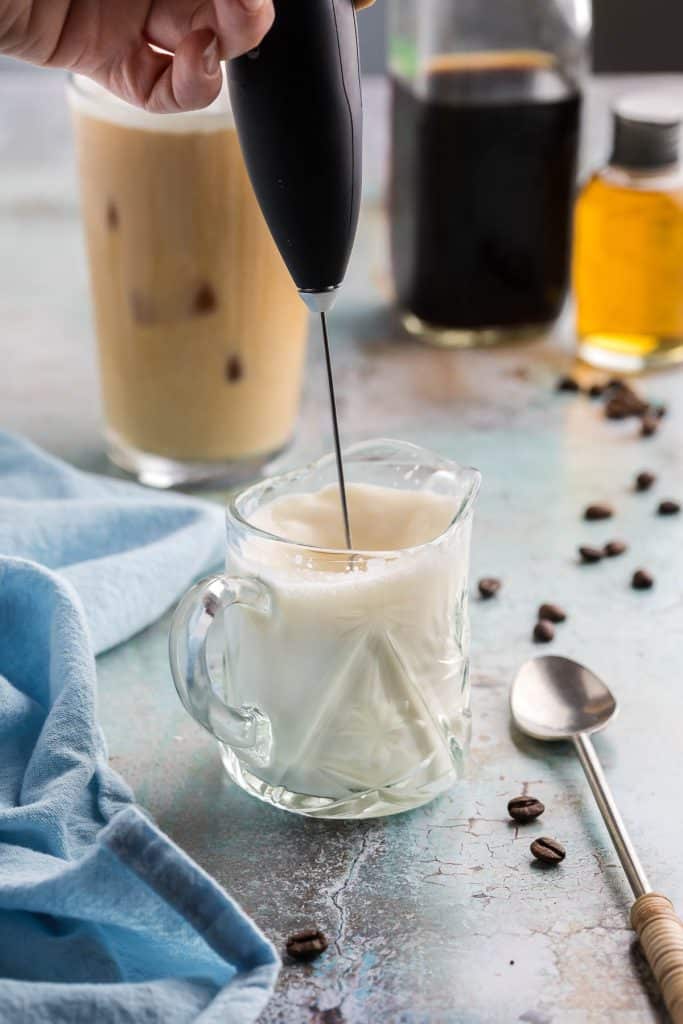 Side shot of a tiny glass jug filled with vanilla sweet cream and being frothed with a frother.
