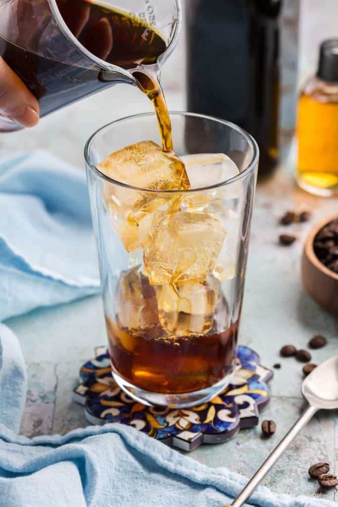 Pouring in the cold brew for the base of the vanilla sweet cream cold brew. A pint glass is filled with ice and the cold brew is cascading over the ice as it is filling up the cup. In the background is a bottle of dark cold brew concentrate, a small bottle of golden vanilla syrup, and a wooden bowl of coffee beans.