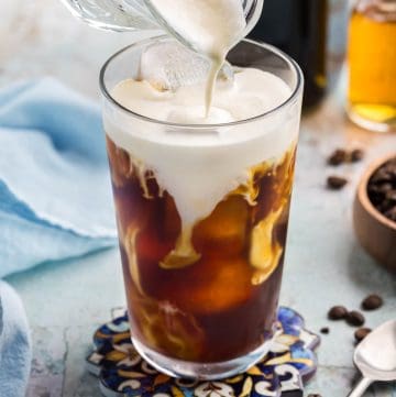 Close up side shot of making a vanilla sweet cream cold brew in a pint glass. The glass is filled with cold brew and ice. Vanilla sweet cream is being poured on to the top surface of the drink, and it is beginning to cascade and swirl with the dark coffee below.