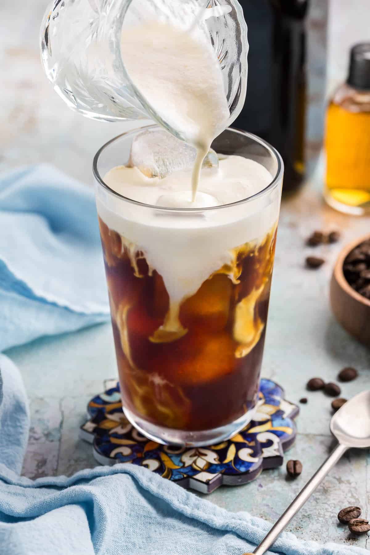 Close up side shot of making a vanilla sweet cream cold brew in a pint glass. The glass is filled with cold brew and ice. Vanilla sweet cream is being poured on to the top surface of the drink, and it is beginning to cascade and swirl with the dark coffee below.