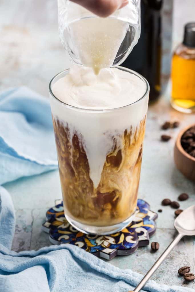 A pint glass filled with vanilla sweet cream cold brew and ice. There is a white frothy top layer of vanilla sweet cream sitting on the top of the drink, and below, most of the cold brew coffee has mixed with vanilla sweet cream and not many swirls are present, but white streaks of cream are still slightly present.