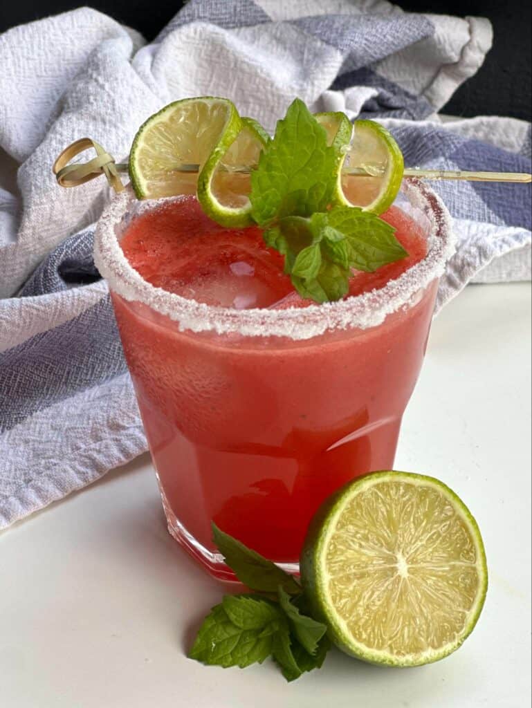 A large glass of watermelon agua fresca. The rim is salted and the red drink is garnished with a splayed out lime wheel on a bamboo cocktail pick. A sliced in half lime is resting up against the glass as well.
