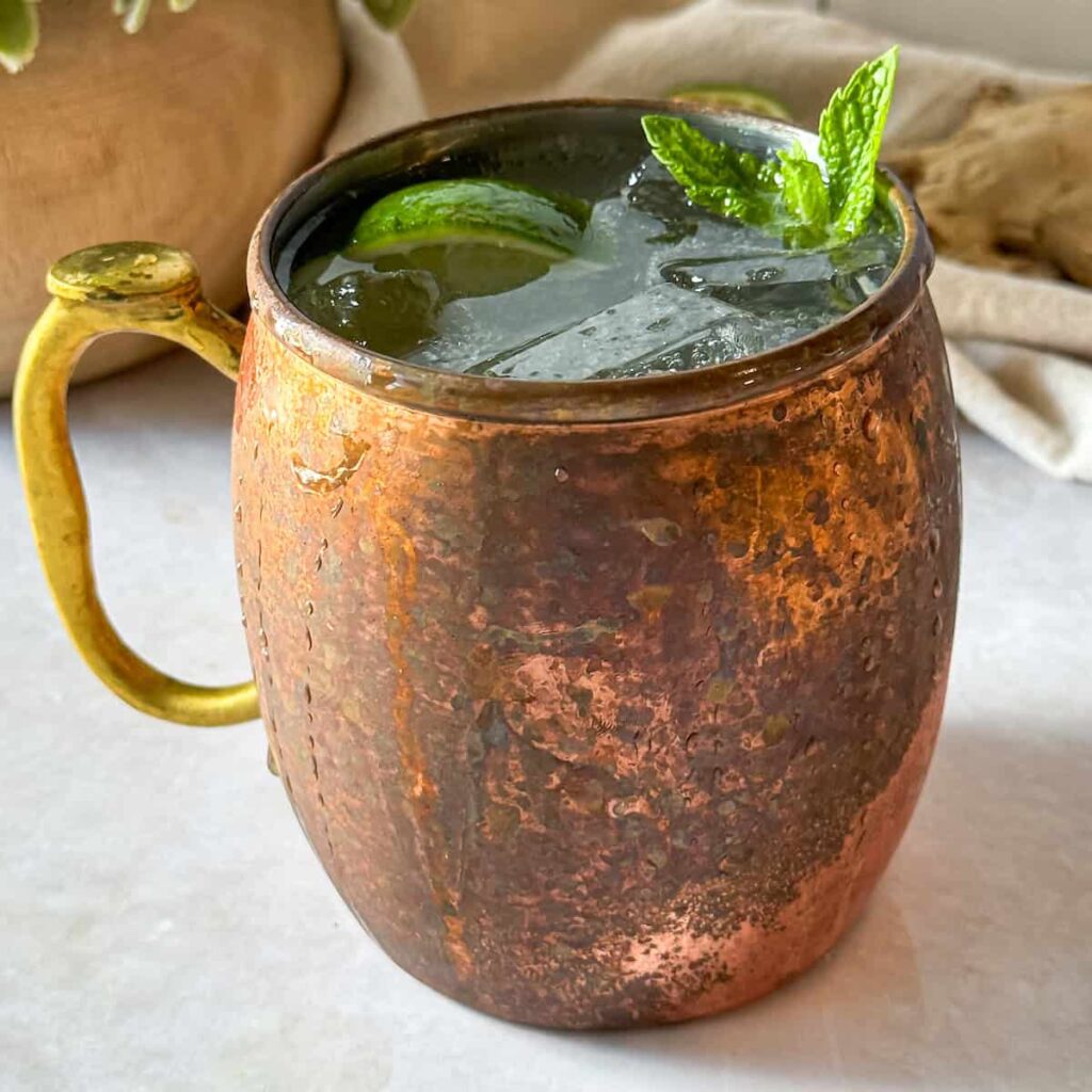 Ginger beer cocktail is a clear fizzy liquid served in a copper mug and it is garnished with lime and fresh mint. The cup is also filled with ice, and condensation is dripping down the copper mug.