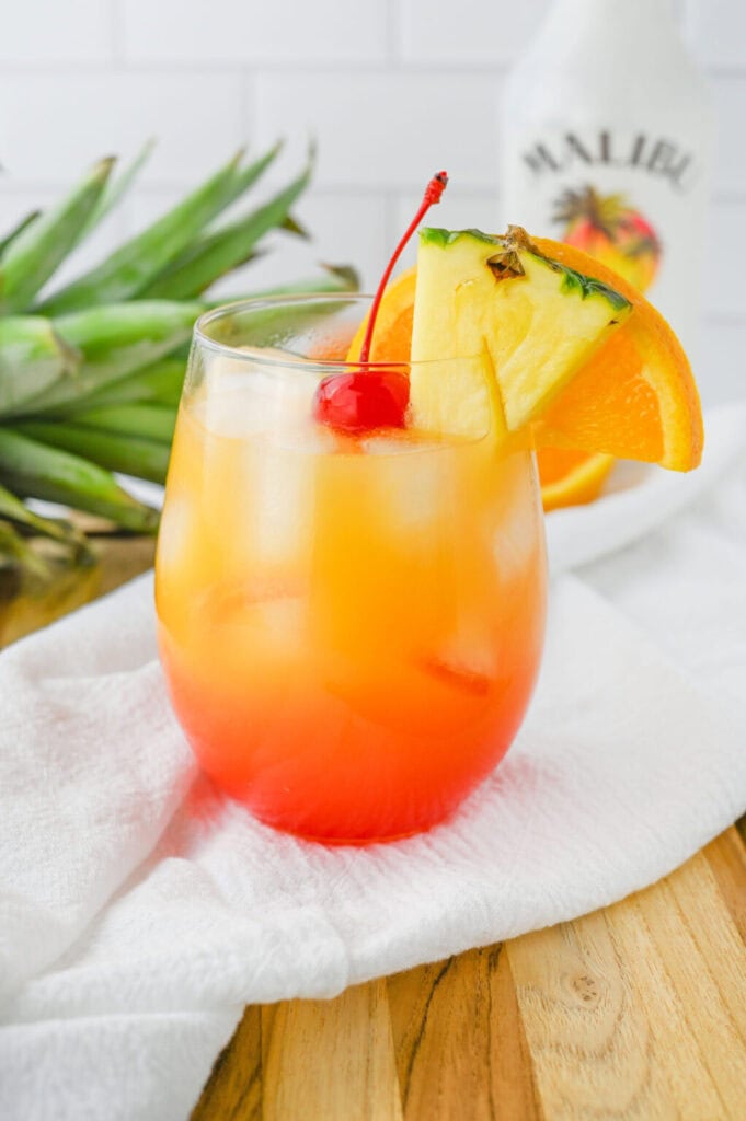 A close up side shot of a malibu sunrise cocktail served in a stemless wine glass. The drink itself is red at the bottom and as you go up the glass the color turns from red to orange. Garnished on top of the ice is a bright red maraschino cherry, and on the edge of the glass is a wedge of fresh pineapple, and a slice of orange.