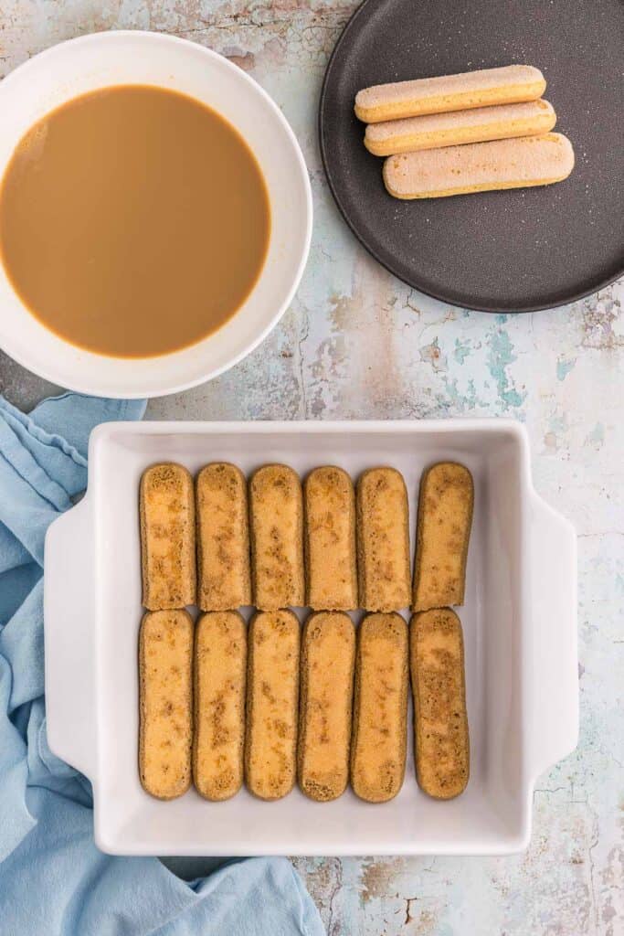 Overhead shot of a white 8x8 casserole dish for Baileys tiramisu layered with two rows of coffee dipped lady fingers.  Above the casserole dish is a shallow bowl filled with the coffee-Irish cream for dipping, and on the right of this bowl is a small black plate with a few dry ladyfingers waiting to be dipped.