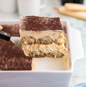 Side shot of a slice of baileys tiramisu being lifted out of a white 8x8 porcelain casserole dish with a small spatula The top is dusted with cocoa powder and the edge shows two layers of espresso soaked lady fingers surrounded by a creamy mascarpone filling. In the background are two small cups of coffee and a bottle of Baileys.