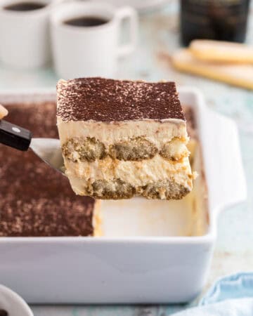 Side shot of a slice of baileys tiramisu being lifted out of a white 8x8 porcelain casserole dish with a small spatula The top is dusted with cocoa powder and the edge shows two layers of espresso soaked lady fingers surrounded by a creamy mascarpone filling. In the background are two small cups of coffee and a bottle of Baileys.