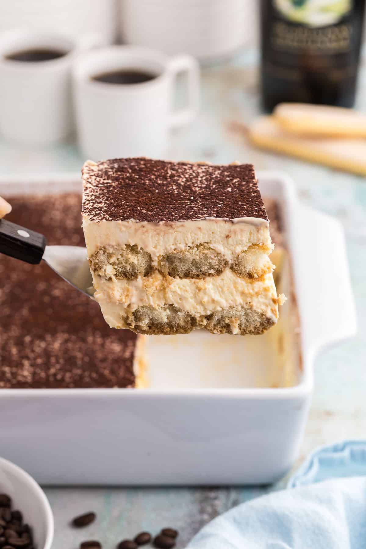 Side shot of a slice of baileys tiramisu being lifted out of a white 8x8 porcelain casserole dish with a small spatula The top is dusted with cocoa powder and the edge shows two layers of espresso soaked lady fingers surrounded by a creamy mascarpone filling. In the background are two small cups of coffee and a bottle of Baileys.