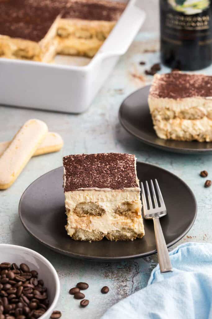 A side shot of a serving of Baileys tiramisu on a small black plate with a fork resting on it.  The two layers of coffee dipped ladyfingers are visible, and the thick creamy mascarpone mixture is topped with a sprinkle of cocoa.  In the foreground a small bowl of coffee beans is peeping in on the left.  In the upper left of the photo is the casserole dish showing some slices taken out.  On the upper right is another black plate with a slice of tiramisu and a bottle of Baileys behind it.