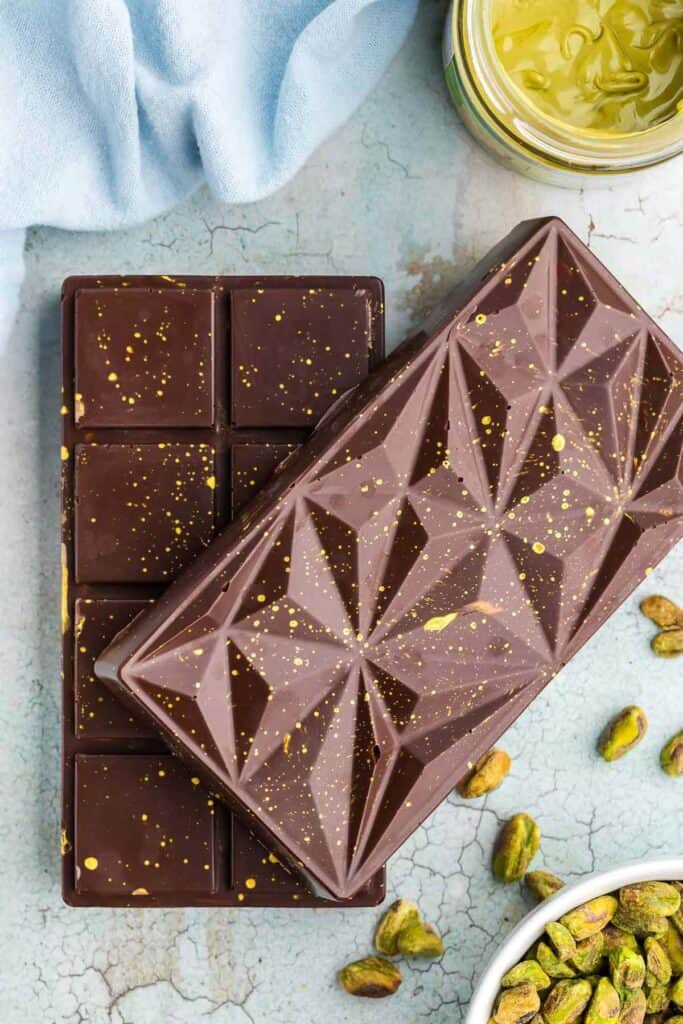 Overhead shot of the two Dubai Chocolate Bars which were made in the chocolate molds.  They are a glossy dark brown with splashes of gold luster on the top.  Fresh pistachios are scattered around the scene.
