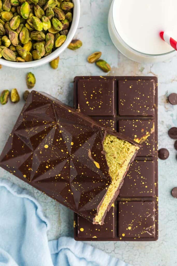 Overhead shot of two Dubai chocolate bars.  The bottom bar is oriented straight up and down and the other bar is laid askew on top and it has been broken op revealing it's green creamy-crunchy pistachio filling.  In the top right corner is a bowl of pistachios.