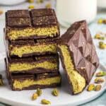 Side shot of four cut open Dubai chocolate bars stacked on top of each other. A fifth bar is laying on its side leaning up against the stack. It too is cut open to reveal a light green filling of pistachio cream and toasted kataifi. In the background is a bowl of green pistachios and a glass of milk. There are also pistachios scattered around the scene.